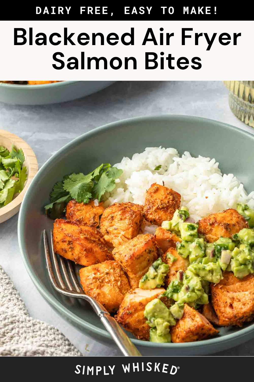 A bowl of white rice and blackened Air Fryer Salmon Bites, topped with creamy avocado cucumber salsa and fresh cilantro, with a fork beside the food. A small bowl of cilantro and a drinking glass are in the background.
