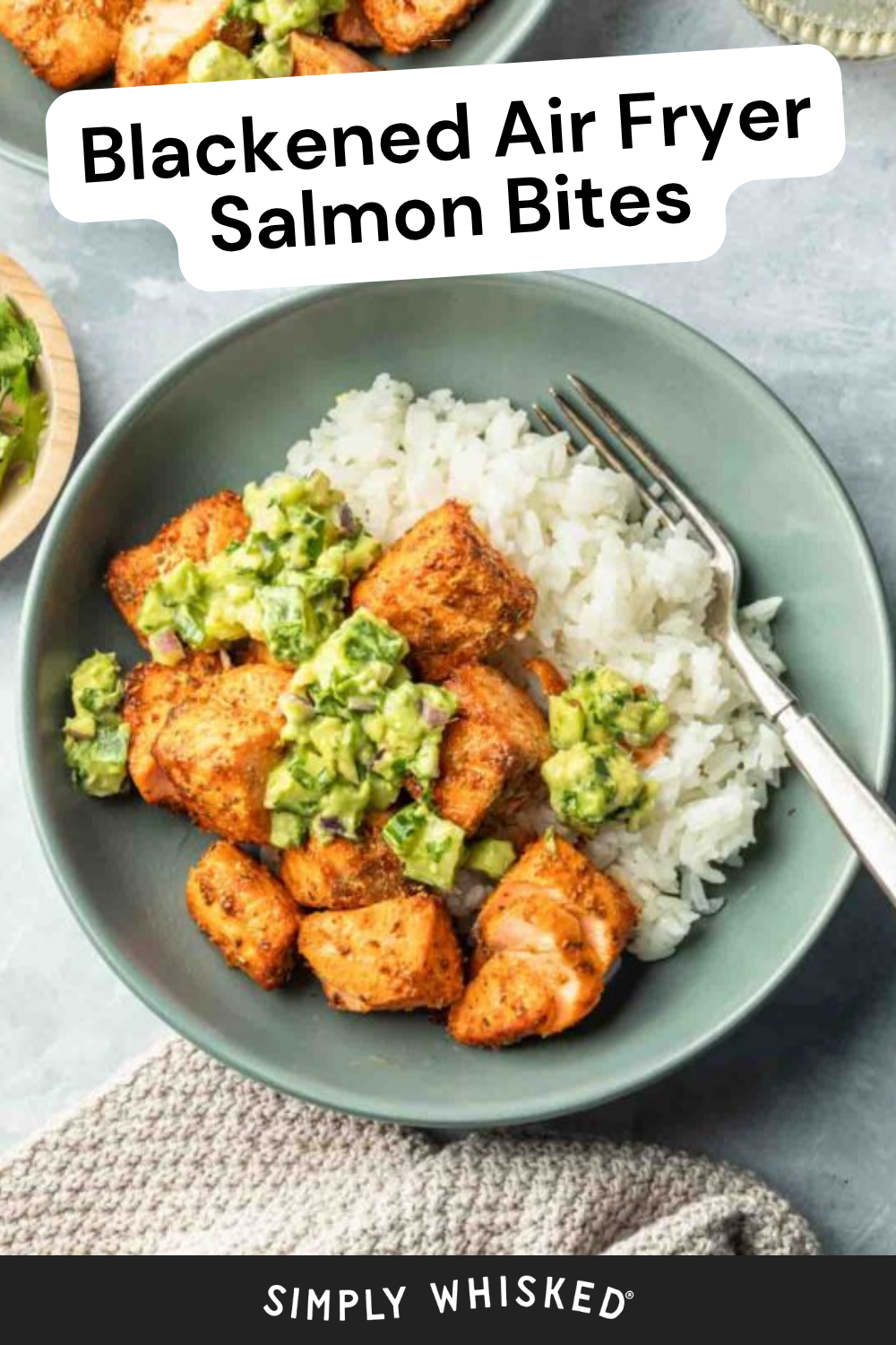 A bowl with white rice and blackened salmon bites topped with a vibrant avocado cucumber salsa, garnished with fresh herbs. A fork rests on the side of the bowl; another bowl and cilantro are partially visible.