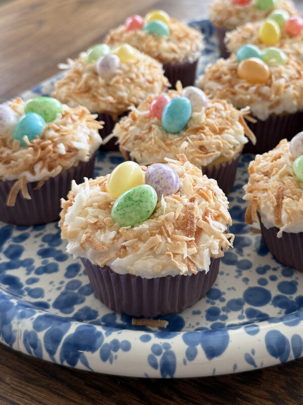 Dairy free coconut cupcakes topped with toasted coconut and colorful candy eggs are displayed on a blue and white patterned plate, resembling Easter nests, on a wooden table.