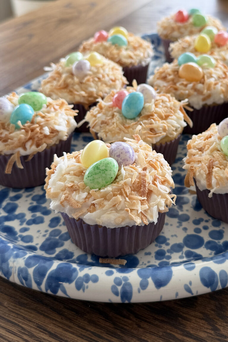 Dairy free coconut cupcakes topped with toasted coconut and colorful candy eggs are displayed on a blue and white patterned plate, resembling Easter nests, on a wooden table.