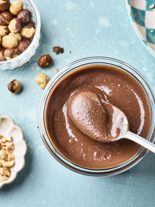 A glass jar of dairy free nutella with a spoon inside sits on a blue surface, surrounded by bowls of whole and chopped hazelnuts.