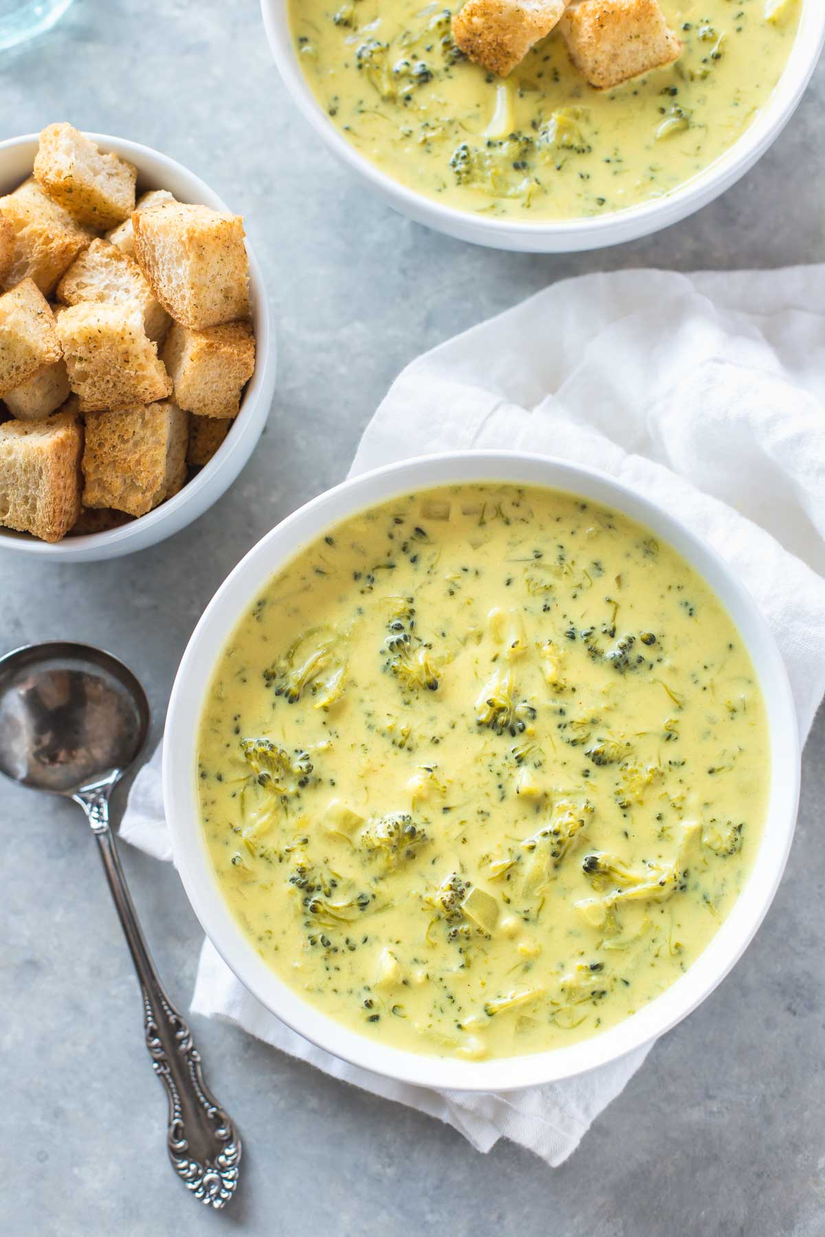 dairy free broccoli cheese soup in a white bowl next to an antique spoon with croutons in a bowl in the background.
