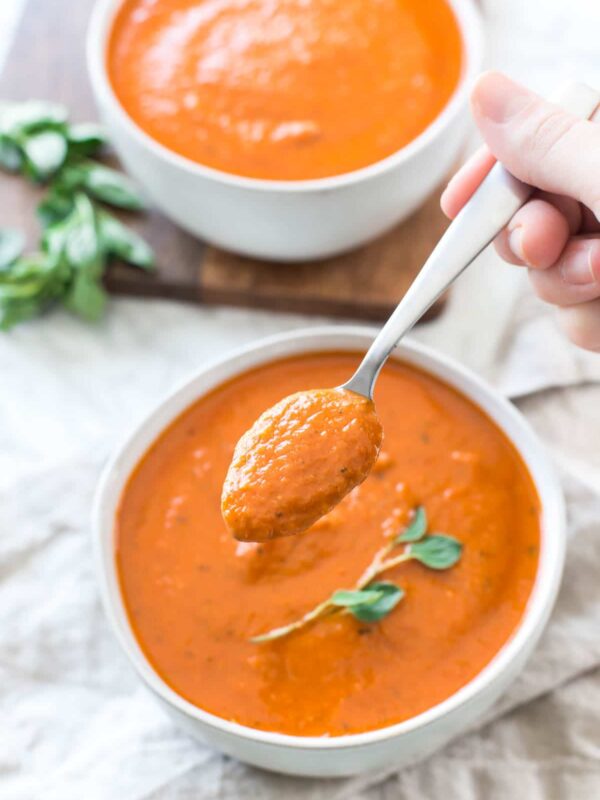 a spoon of creamy dairy free tomato soup above a full bowl on a tan linen napkin with fresh oregano garnish and a second bowl of soup in the background