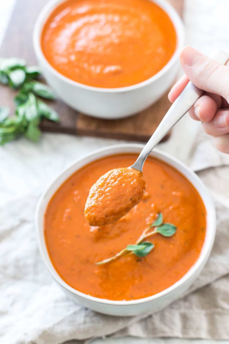 a spoon of creamy dairy free tomato soup above a full bowl on a tan linen napkin with fresh oregano garnish and a second bowl of soup in the background