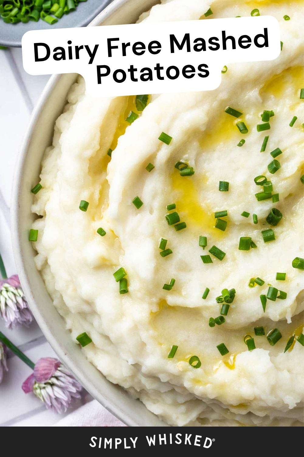 A bowl of creamy dairy free mashed potatoes topped with chopped chives and a drizzle of olive oil. Text overlay reads Dairy Free Mashed Potatoes. Decorative flowers and chives are visible beside the bowl, highlighting this delicious vegan mashed potatoes option.