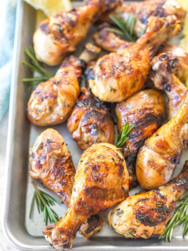 A baking tray filled with Grilled Chicken Drumsticks, garnished with sprigs of fresh rosemary and lemon wedges in the corners.