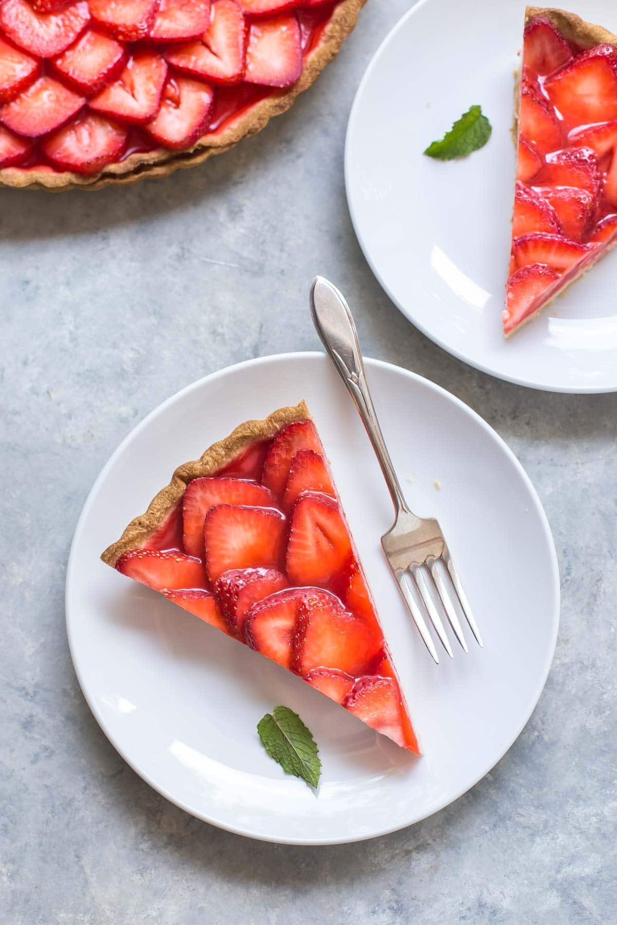 sliced strawberry tart on a dessert plate with a small mint leaf and fork, another serving and the rest of the tart in the background
