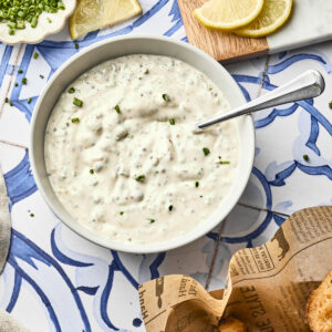 A bowl of creamy homemade tartar sauce with chopped chives sits on a blue and white tiled surface, surrounded by lemon wedges, fresh herbs, and a basket of fried fish pieces on newspaper-style paper—a classic tartar sauce recipe scene.