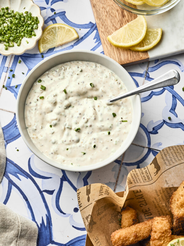 A bowl of creamy homemade tartar sauce with chopped chives sits on a blue and white tiled surface, surrounded by lemon wedges, fresh herbs, and a basket of fried fish pieces on newspaper-style paper—a classic tartar sauce recipe scene.