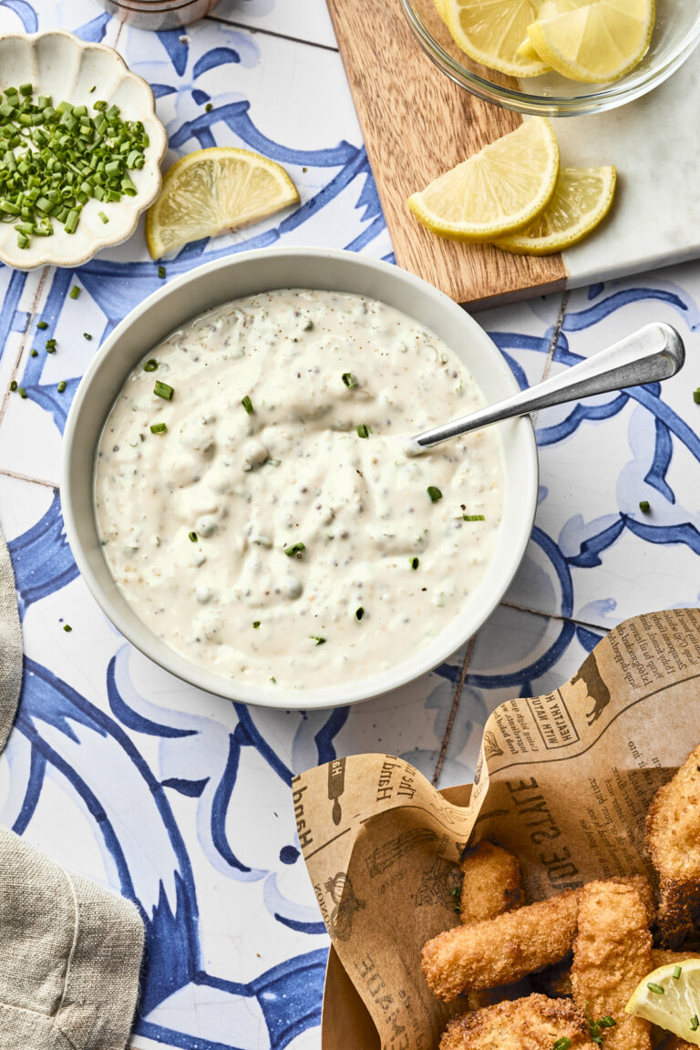 A bowl of creamy homemade tartar sauce with chopped chives sits on a blue and white tiled surface, surrounded by lemon wedges, fresh herbs, and a basket of fried fish pieces on newspaper-style paper—a classic tartar sauce recipe scene.