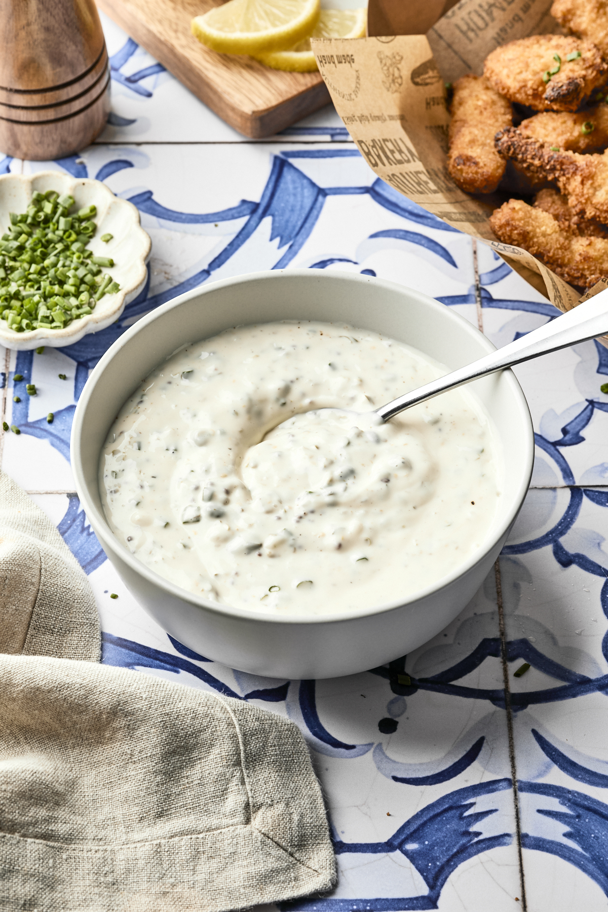 A white bowl filled with homemade tartar sauce and a spoon sits on a decorative blue and white tile. Nearby are chopped chives, a beige napkin, lemon wedges, and breaded fried food in a basket—perfect for trying your next tartar sauce recipe.
