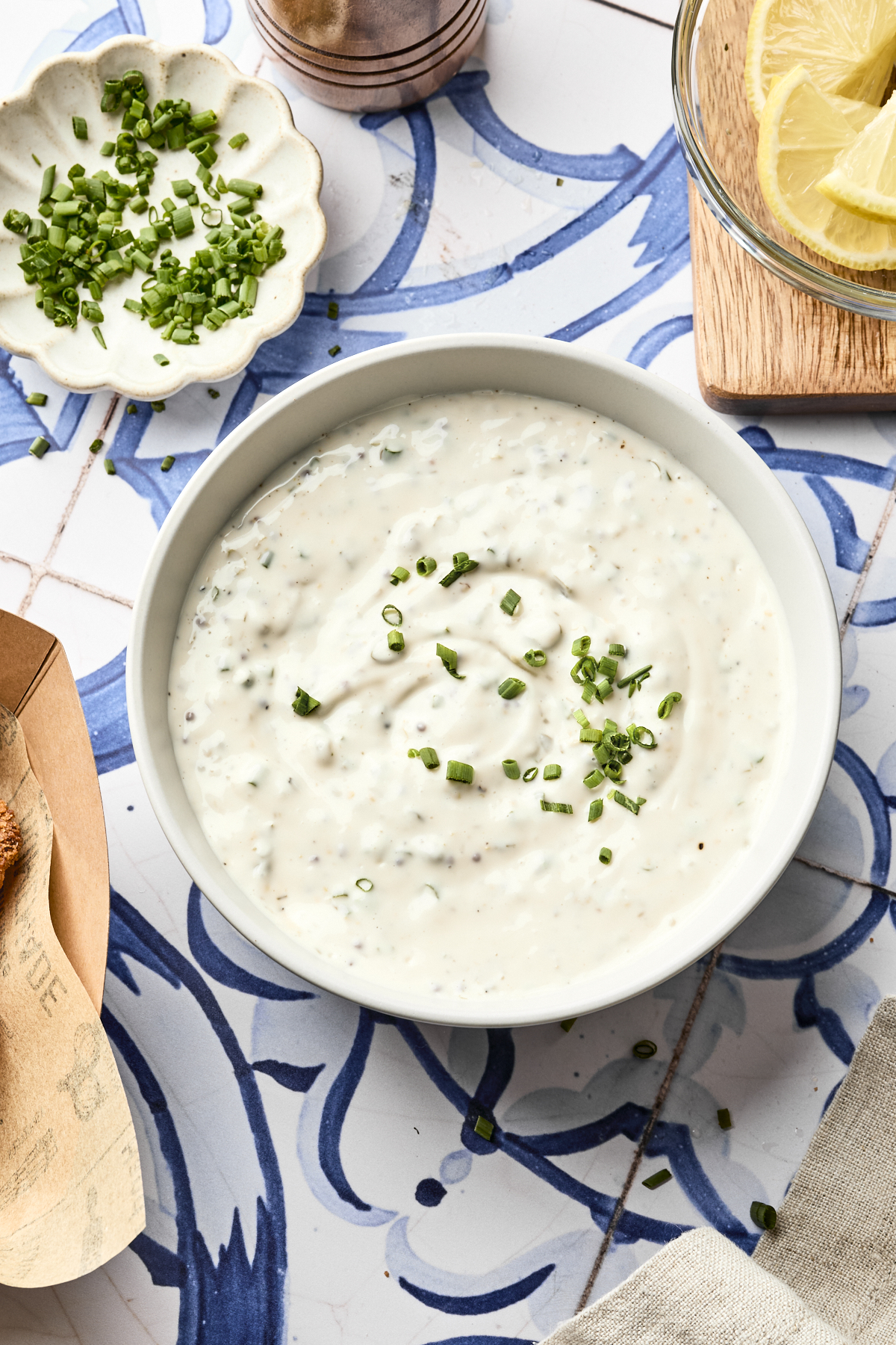 A bowl of creamy homemade tartar sauce topped with chopped chives sits on a blue and white tiled surface. Nearby are a dish of more chives, lemon slices, and part of a wooden utensil. Perfect for your favorite tartar sauce recipe!.