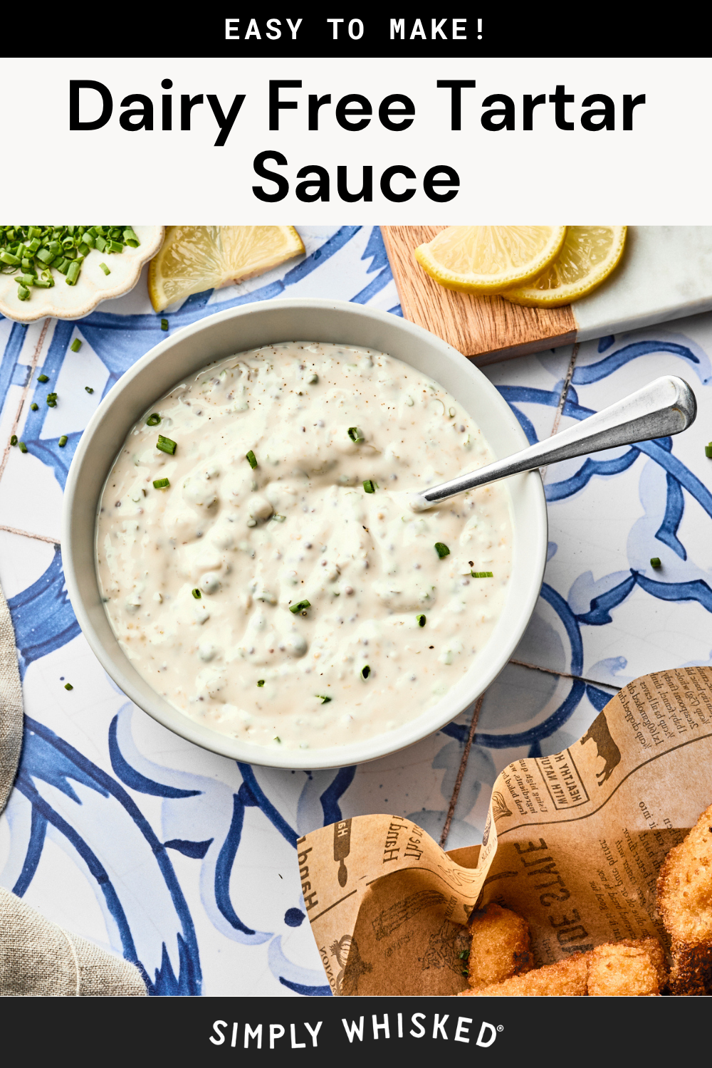 A bowl of dairy-free tartar sauce with a spoon, garnished with chopped herbs. Lemon wedges, chopped chives, and breaded food are nearby on a tiled surface. Text: Dairy Free Tartar Sauce. Simply Whisked. Discover this easy homemade tartar sauce for a delicious twist on the classic tartar sauce recipe.