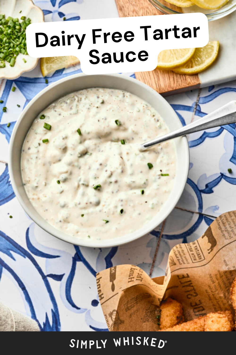 A bowl of homemade tartar sauce with chopped chives, lemon slices, and a fork on a blue and white patterned surface. Some breaded food sits nearby on brown paper. Text reads Dairy Free Tartar Sauce—perfect for your favorite tartar sauce recipe.