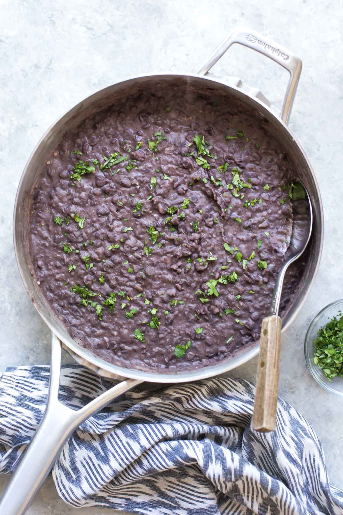 Aerial view of a skillet with refried black beans topped with fresh cilantro, black and white towel under the handle and a small bowl of cilantro on the side.