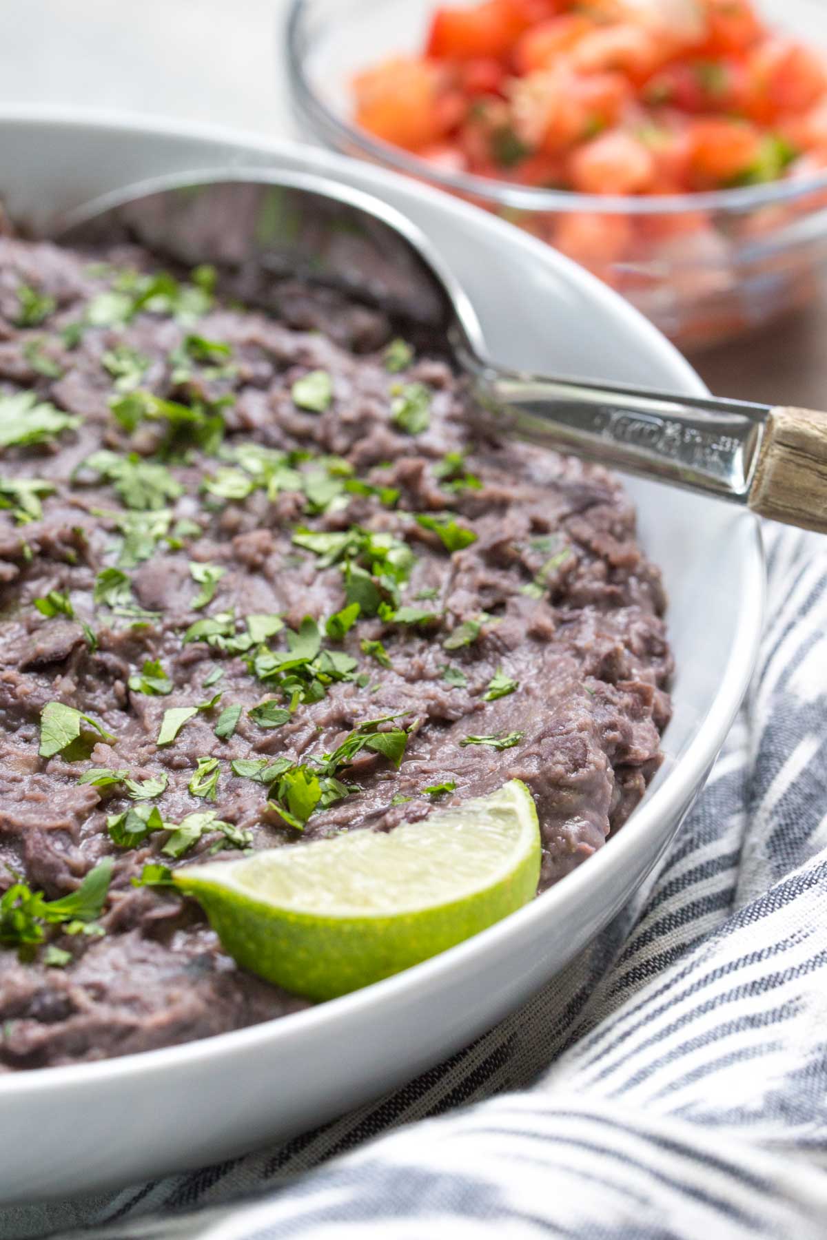 closeup on refried black beans topped with cilantro and a lime wedge in a white bowl with a metal spoon with wooden handle