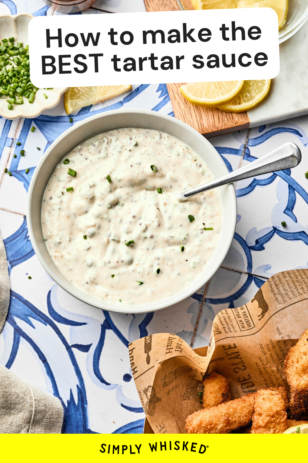 A bowl of homemade tartar sauce with chopped herbs on top sits on a blue and white patterned surface. A spoon is in the bowl, and fish sticks, lemon slices, and green onions are nearby. Text reads: “How to make the BEST tartar sauce recipe.”.