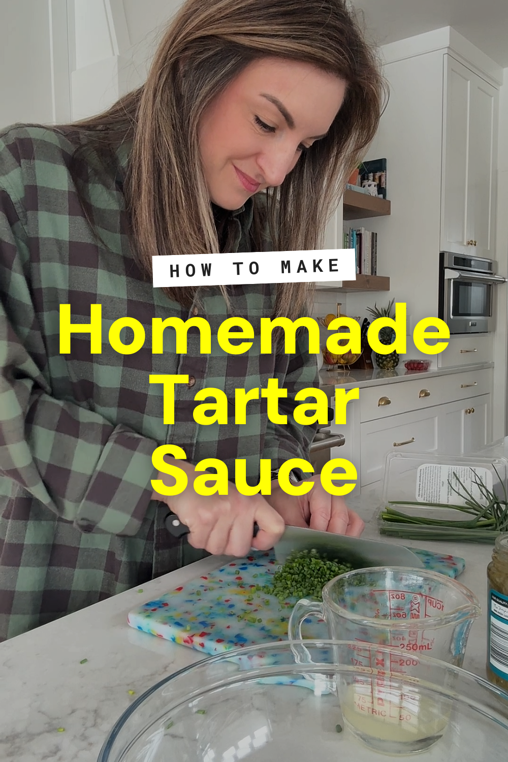 A woman in a plaid shirt chops herbs on a colorful cutting board in a modern kitchen. Text overlay reads “How to make Homemade Tartar Sauce.” Measuring cups and tartar sauce recipe ingredients are on the counter.