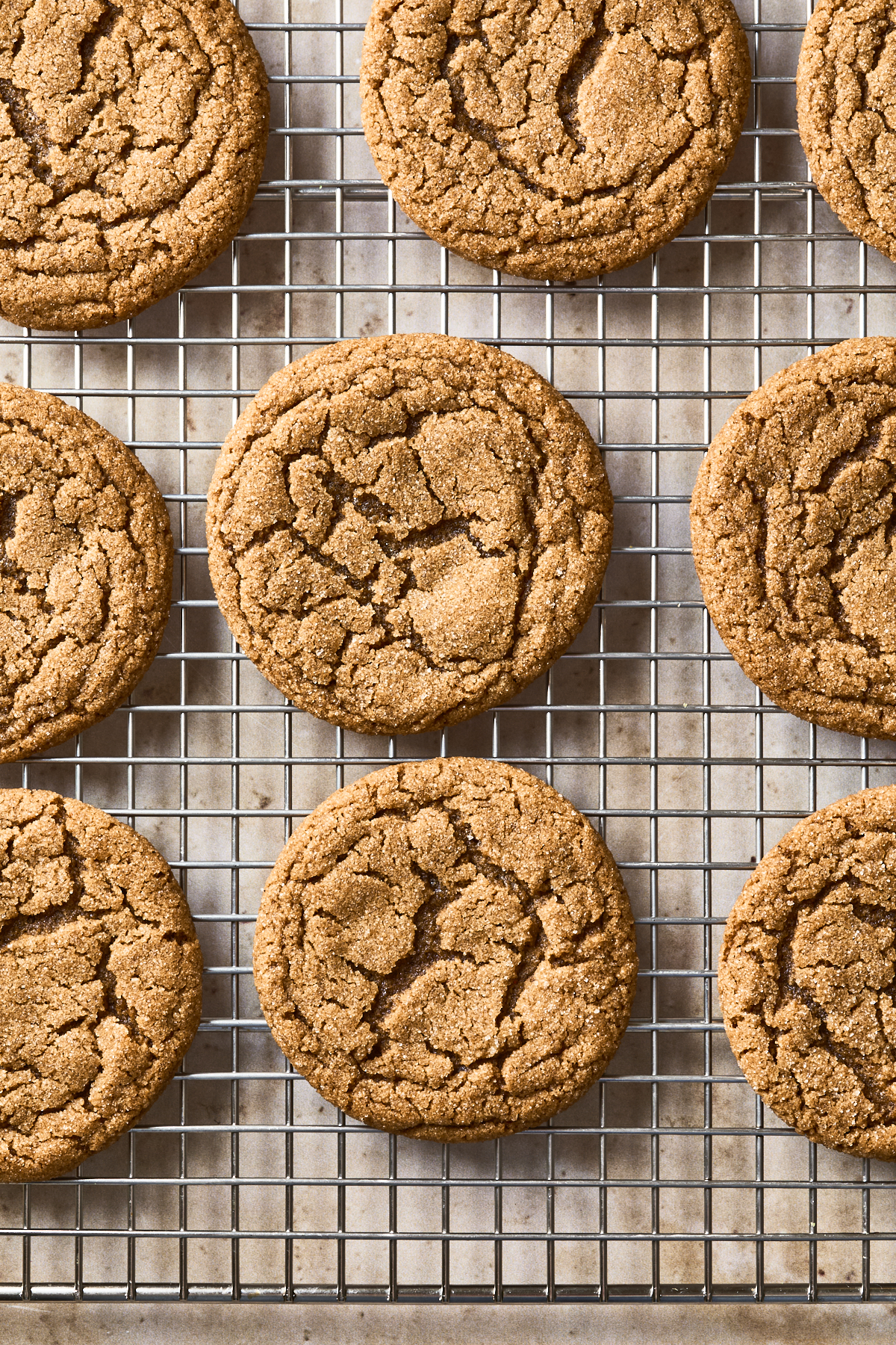 Nine golden brown soft gingersnap cookies with crackled tops cool on a wire rack, arranged in neat rows over a light-colored surface.