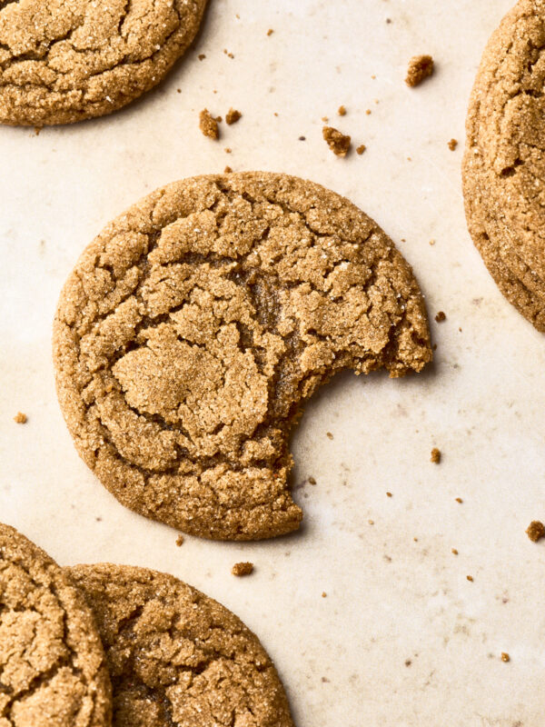 A close-up of several cracked soft gingersnap cookies on parchment paper, with one cookie in the center missing a bite and surrounded by crumbs.