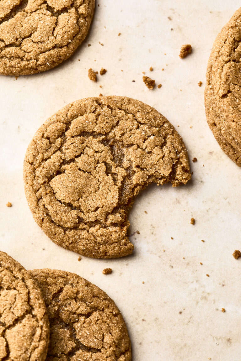 A close-up of several cracked soft gingersnap cookies on parchment paper, with one cookie in the center missing a bite and surrounded by crumbs.