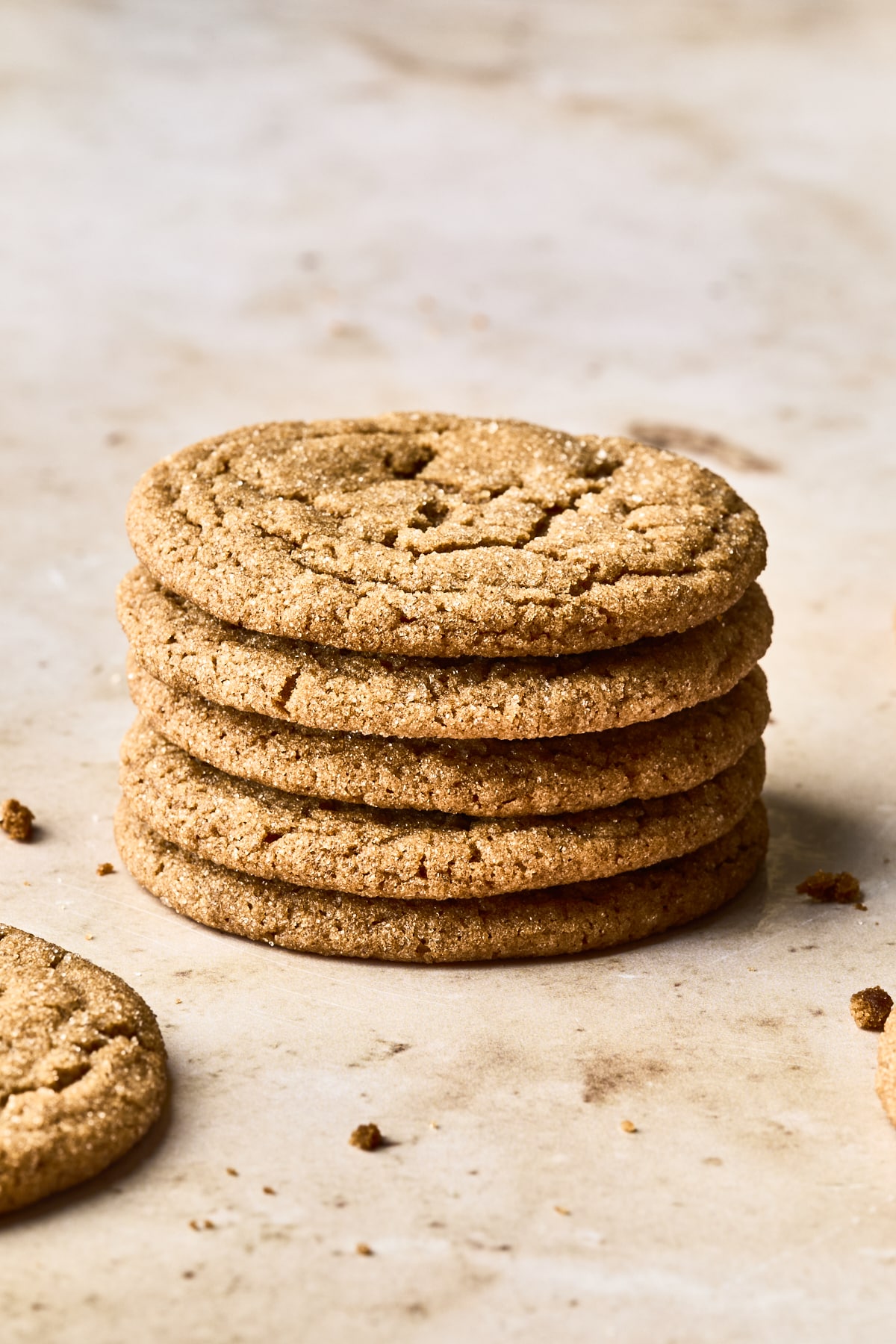 A stack of five golden-brown soft gingersnap cookies sits on a light, textured surface, with another cookie and a few crumbs scattered nearby.
