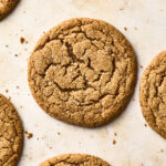 A close-up view of several round, golden-brown soft gingersnap cookies with a cracked surface, arranged on a light-colored baking sheet.