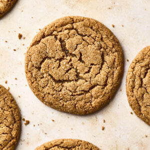 A close-up view of several round, golden-brown soft gingersnap cookies with a cracked surface, arranged on a light-colored baking sheet.