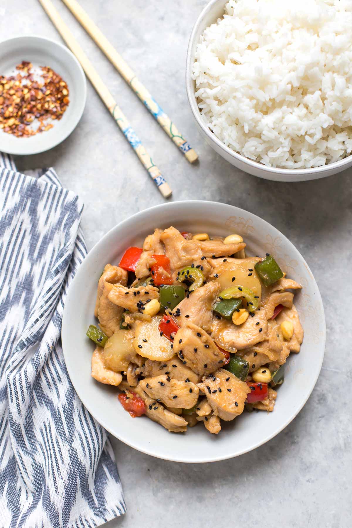 kung pao chicken on a small plate next to a bowl of rice and a small bowl of red pepper flakes