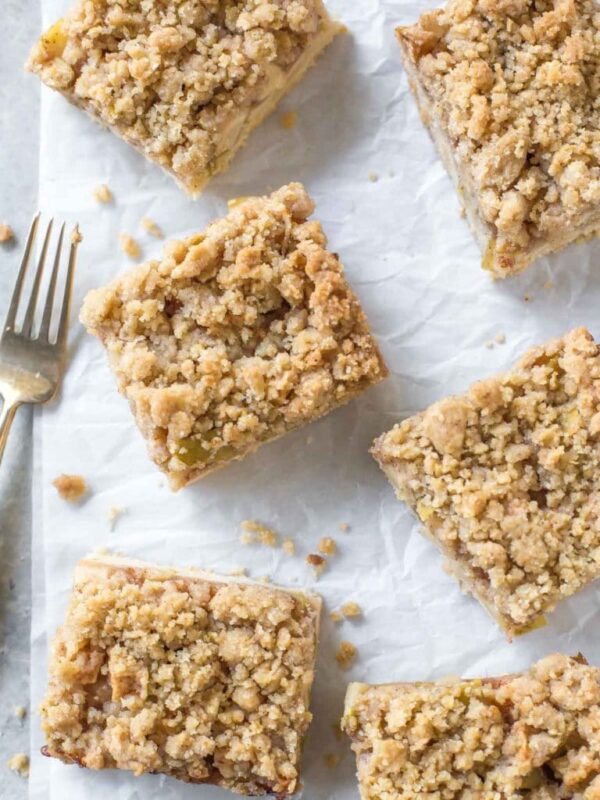 close up overhead view of 6 apple pie bars with crumb topping on a crinkled piece of white parchment paper, a small gold fork with wooden handle next to them