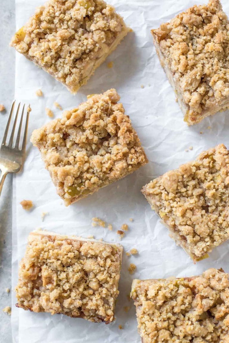close up overhead view of 6 apple pie bars with crumb topping on a crinkled piece of white parchment paper, a small gold fork with wooden handle next to them