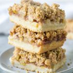 three apple pie bars stacked on a small gray plate on a gray surface with an apple crumb bar in the background and a gold fork with wooden handle in the foreground - with text overlay optimized for Pinterest
