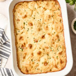 A baked dairy free scalloped potatoes gratin with a golden, bubbly top is in a white rectangular baking dish, garnished with chopped herbs. A striped kitchen towel, salt and pepper shakers, and fresh parsley are nearby.