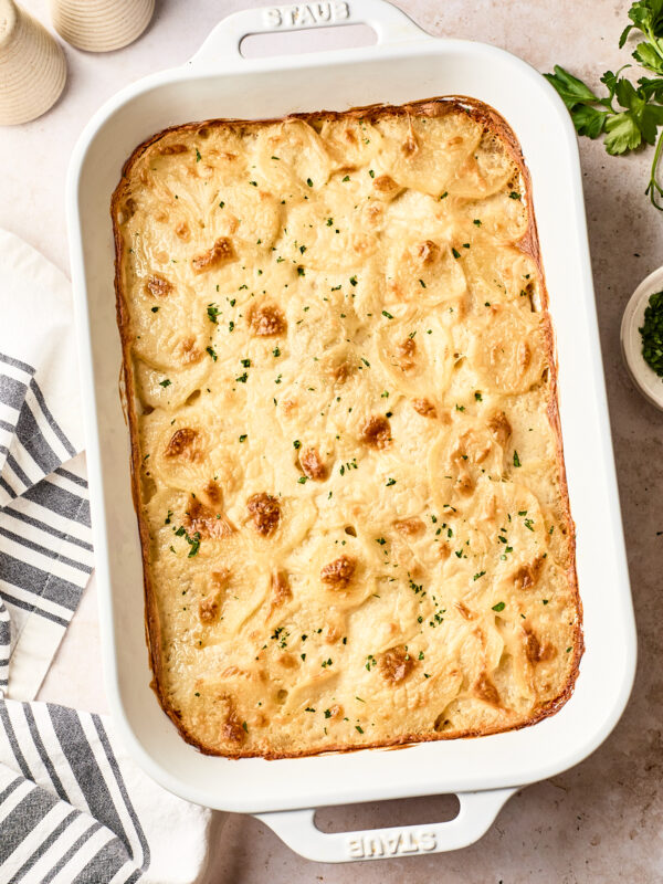 A baked dairy free scalloped potatoes gratin with a golden, bubbly top is in a white rectangular baking dish, garnished with chopped herbs. A striped kitchen towel, salt and pepper shakers, and fresh parsley are nearby.