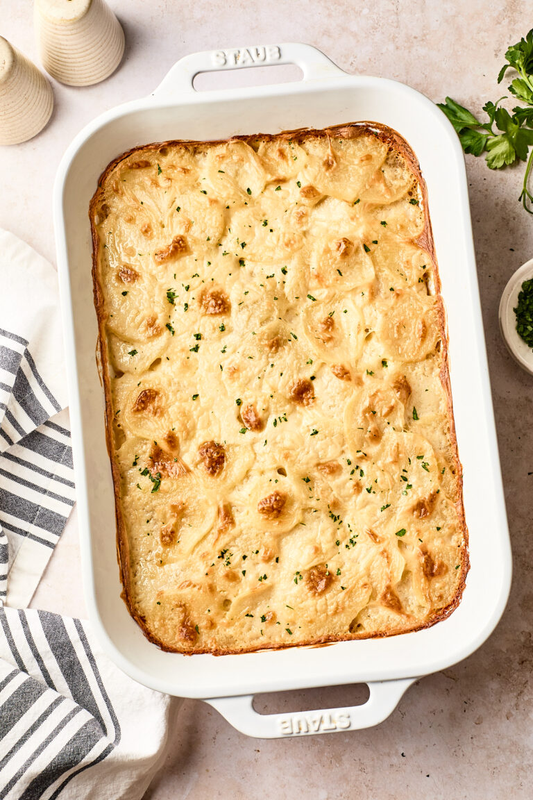 A baked dairy free scalloped potatoes gratin with a golden, bubbly top is in a white rectangular baking dish, garnished with chopped herbs. A striped kitchen towel, salt and pepper shakers, and fresh parsley are nearby.