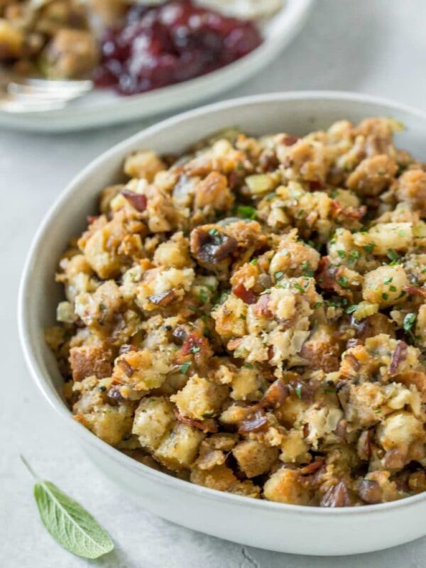 close up of pancetta chestnut stuffing in a bowl with next to two plates with stuffing, turkey, gravy and cranberry sauce, on a gray background with fresh sage leaves and a linen napkin to the side