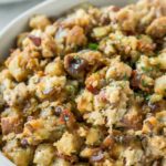 close up of pancetta chestnut stuffing in a bowl with next to two plates with stuffing, turkey, gravy and cranberry sauce, on a gray background with fresh sage leaves and a linen napkin to the side, optimized for Pinterest with a text overlay