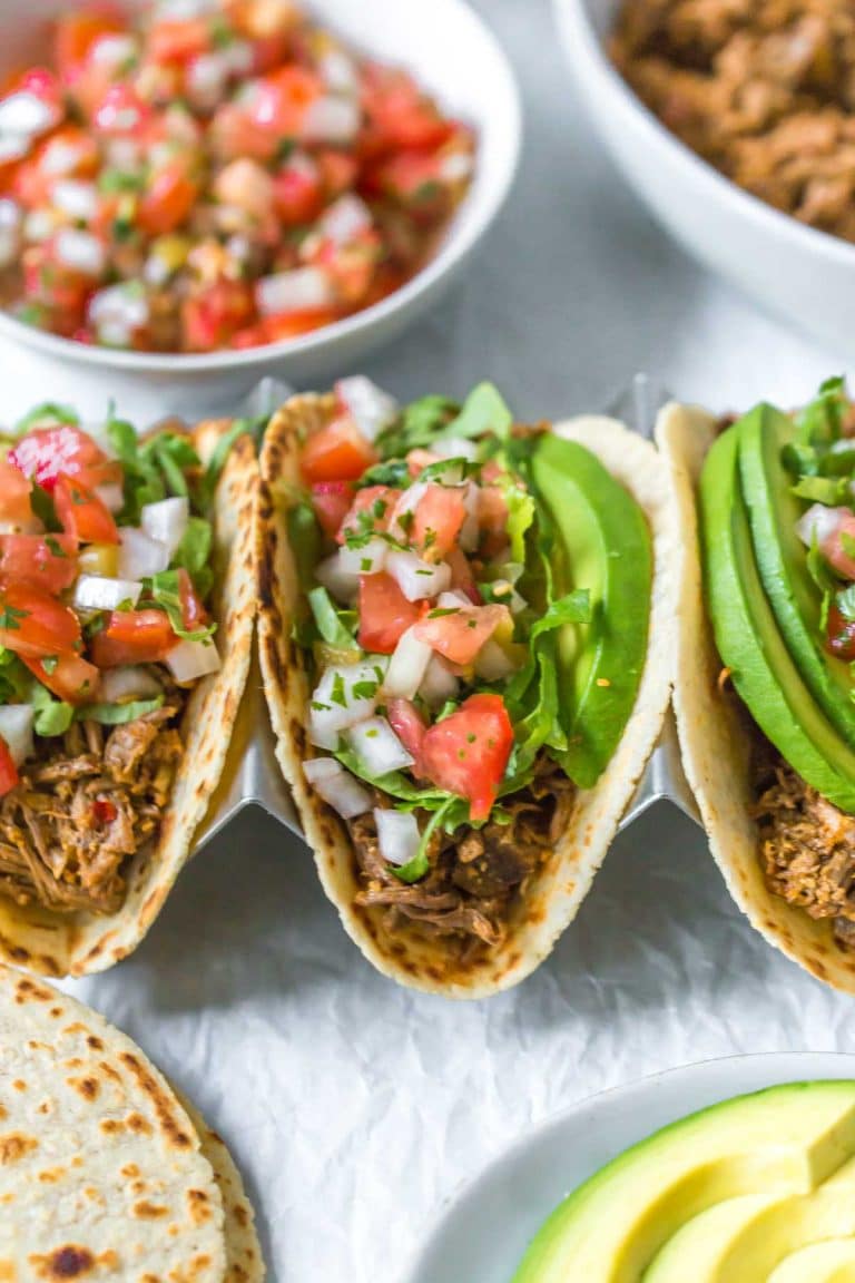closeup view of slow cooker beef tacos topped with shredded lettuce, pico de gallo and avocado slices, surrounded by taco toppings in bowls, halved avocados and tortillas