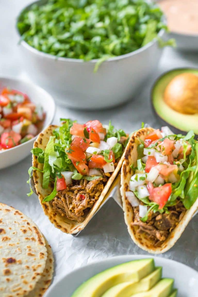 45 degree angle view of slow cooker beef tacos topped with shredded lettuce, pico de gallo and avocado slices, surrounded by taco toppings in bowls, halved avocados and tortillas