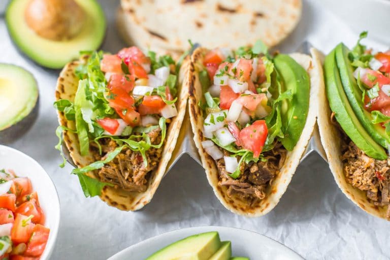45 degree angle view of slow cooker beef tacos topped with shredded lettuce, pico de gallo and avocado slices, surrounded by taco toppings in bowls, halved avocados and tortillas
