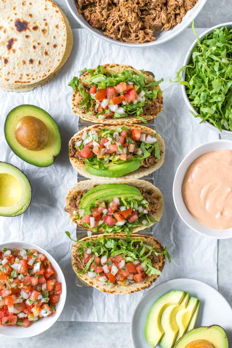 slow cooker beef tacos topped with shredded lettuce, pico de gallo and avocado slices, surrounded by taco toppings in bowls, halved avocados and tortillas