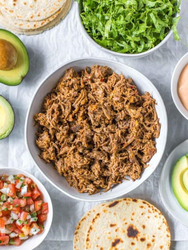 overhead view of slow cooker shredded beef taco meat in a bowl, surrounded by taco toppings in bowls, tortillas and avocados