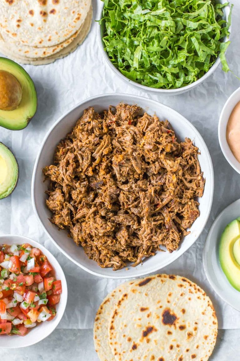 overhead view of slow cooker shredded beef taco meat in a bowl, surrounded by taco toppings in bowls, tortillas and avocados