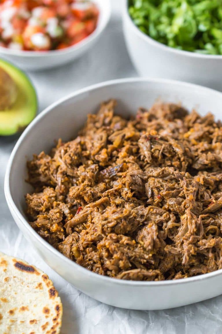 closeup of slow cooker shredded beef taco meat in a bowl, surrounded by taco toppings in bowls, tortillas and avocados