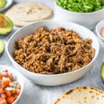 slow cooker shredded beef taco meat in a bowl, surrounded by taco toppings in bowls, tortillas and avocados