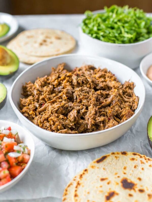 slow cooker shredded beef taco meat in a bowl, surrounded by taco toppings in bowls, tortillas and avocados