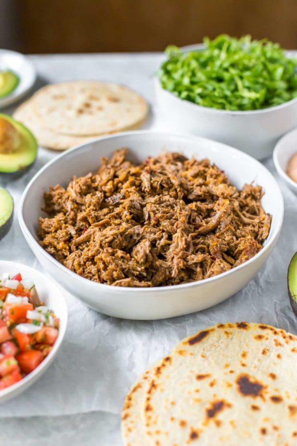 slow cooker shredded beef taco meat in a bowl, surrounded by taco toppings in bowls, tortillas and avocados