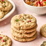 A stack of four fruitcake cookies with colorful candied fruit pieces sits on a pink surface. More cookies and a bowl of mixed candied fruit are in the background.