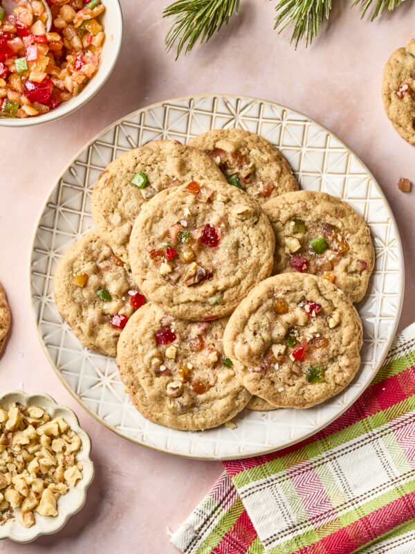 A plate of fruitcake cookies with colorful candied fruit and chopped nuts sits on a pink surface, surrounded by a bowl of candied fruit, a dish of nuts, pine branches, and a red and green plaid napkin.