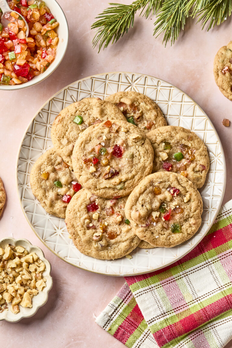 A plate of fruitcake cookies with colorful candied fruit and chopped nuts sits on a pink surface, surrounded by a bowl of candied fruit, a dish of nuts, pine branches, and a red and green plaid napkin.